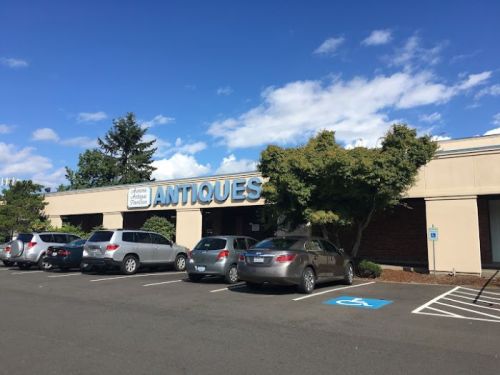 A building with a sign reading "ANTIQUES" and several parked cars in front under a blue sky with clouds.
