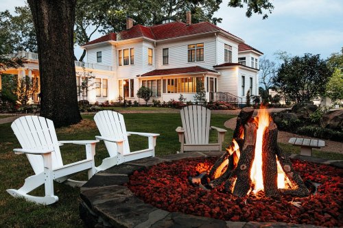 A cozy outdoor scene featuring a fire pit with flames, surrounded by white chairs and a beautiful house in the background.