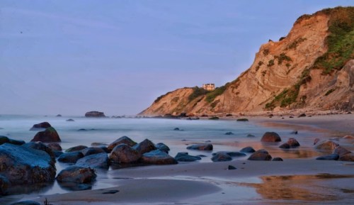 A serene beach scene with smooth waves, rocky shore, and a distant cliffside house under a soft twilight sky.