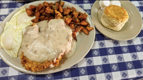 A plate of fried chicken with gravy, fried potatoes, two eggs, and a biscuit on the side, all on a checkered tablecloth.