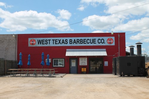 Exterior of West Texas Barbecue Co. featuring a red building, picnic tables, and a smoker. Blue sky with clouds above.