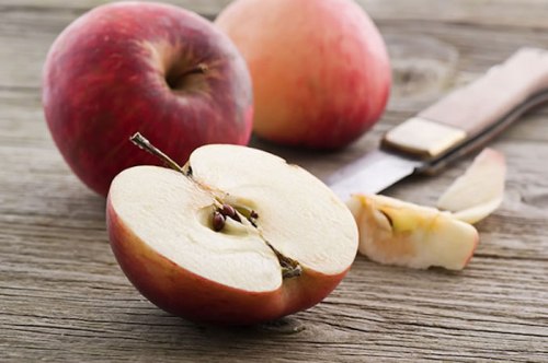 A sliced red apple on a wooden surface, with whole apples and a knife in the background.