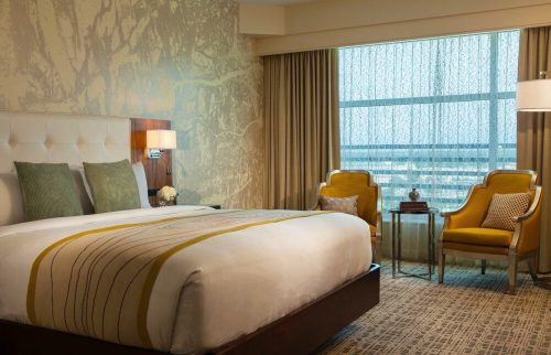 A modern hotel room featuring a king bed, two chairs, a side table, and large windows with sheer curtains.