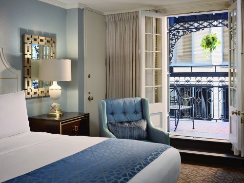 A cozy hotel room with a bed, lamp, and armchair, featuring a balcony with a view of a wrought-iron railing and greenery.