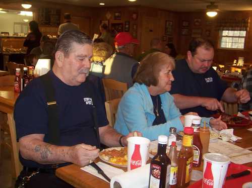 Three people enjoying a meal at a restaurant, with condiments and drinks on the table. Casual dining atmosphere.