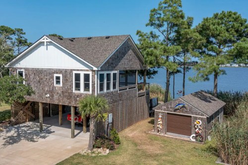 A charming waterfront house with a wooden exterior, surrounded by trees and a small shed, overlooking a calm lake.
