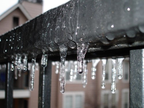Icicles hanging from a metal railing, with droplets of water glistening in the light. Background shows a blurred building.