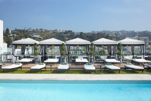 A rooftop pool area with lounge chairs and cabanas, surrounded by greenery and a scenic hillside view.