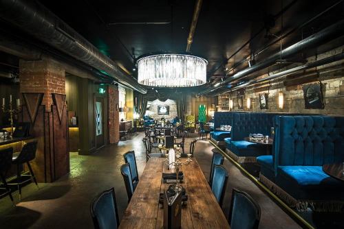 A stylish restaurant interior featuring blue velvet seating, a large chandelier, and rustic wooden tables.