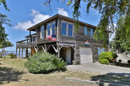 A wooden house on stilts with an American flag, surrounded by greenery and a clear blue sky.