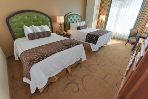 A hotel room featuring two beds with green headboards, decorative bedding, and large windows letting in natural light.