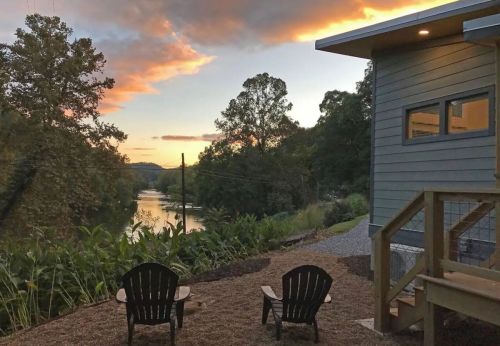 A serene riverside view at sunset, featuring two chairs and a modern house surrounded by trees and greenery.
