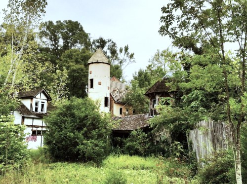 Abandoned buildings surrounded by overgrown greenery, featuring a tower and a rustic, weathered appearance.