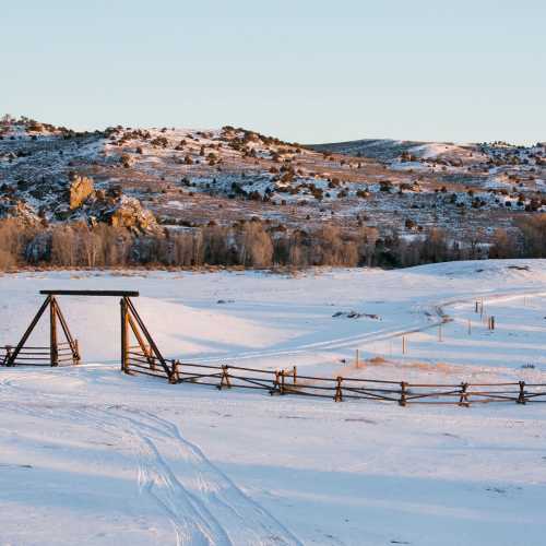 Snow-covered landscape with a wooden fence and bridge, surrounded by hills and trees under a clear sky.