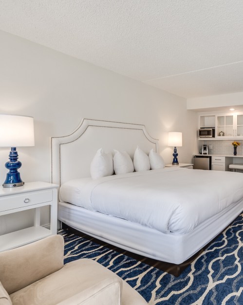 A modern hotel room featuring a large bed, nightstands, lamps, and a kitchenette with a cozy seating area.
