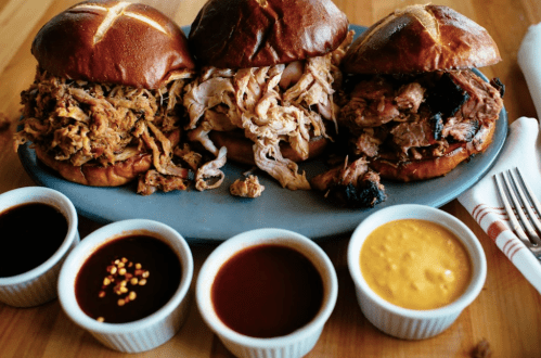 Three pretzel buns filled with pulled meats, served on a platter with four small bowls of different sauces.