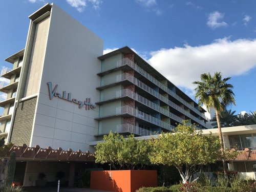 Modern hotel building with balconies, palm trees, and a blue sky in the background. Sign reads "Valley Ho."