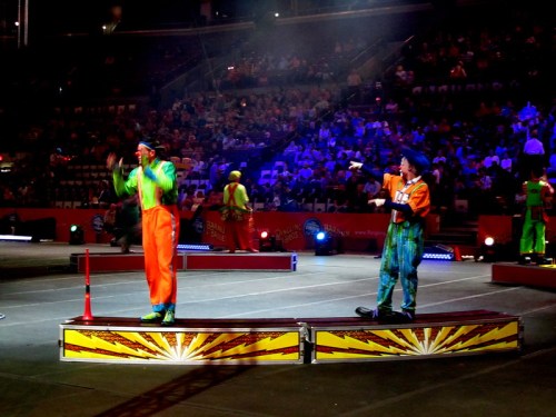 Two clowns perform on stage in a circus, with a cheering audience in the background and colorful lighting.