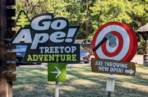 Signage for "Go Ape! Treetop Adventure" with an arrow and a sign for "Axe Throwing Now Open!" in a park setting.