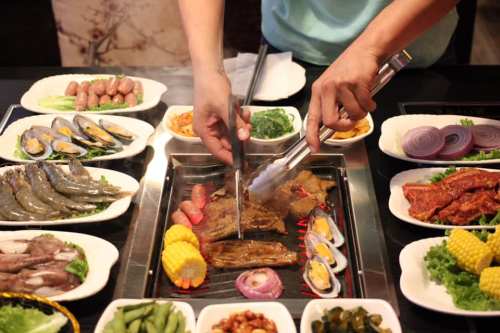 A person grilling various meats and seafood at a table surrounded by fresh vegetables and side dishes.