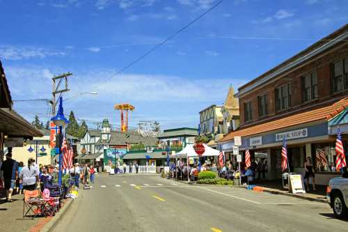 A lively street scene with shops, flags, and people enjoying a sunny day at a festival in a small town.