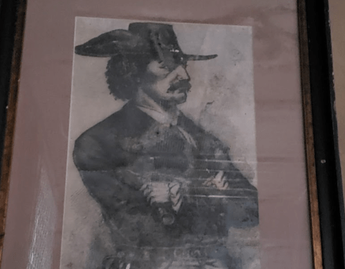 A vintage portrait of a man with a mustache, wearing a hat and coat, arms crossed, displayed in a frame.