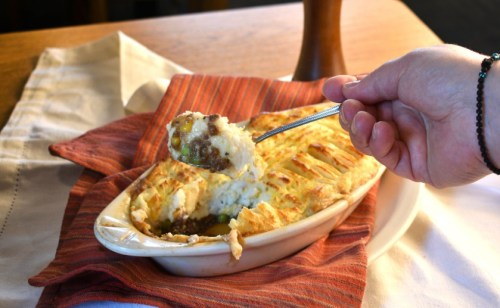 A hand holding a spoonful of shepherd's pie, with a baked dish on a table, surrounded by a striped napkin.