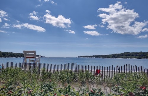 A serene beach scene with a lifeguard chair, blue water, and fluffy clouds under a bright sky, framed by a wooden fence.