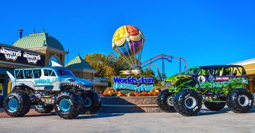 Two colorful monster trucks are parked in front of a theme park entrance with a large balloon and roller coaster in the background.