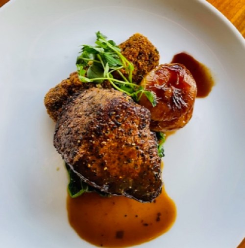 A plated dish featuring a seasoned meat cut, crispy sides, a roasted tomato, and a drizzle of sauce, garnished with greens.