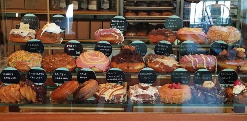 A display case filled with various colorful donuts, each labeled with names and prices.