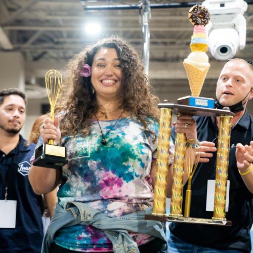 A smiling woman holds a trophy and a whisk, celebrating her win at an ice cream competition.