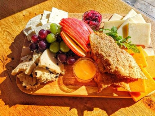 A wooden platter with assorted cheeses, grapes, apple slices, crackers, and a small bowl of dipping sauce.