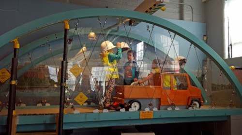 Children play in a colorful interactive exhibit featuring a toy truck and safety gear in a museum setting.