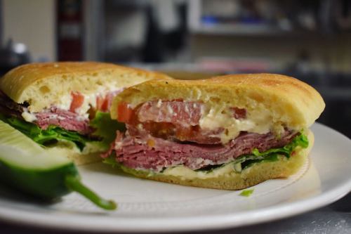 A close-up of a sandwich with layers of meat, lettuce, tomato, and creamy dressing on a toasted bun, served with a pickle.