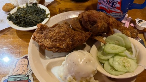 A plate of fried chicken, mashed potatoes with gravy, cucumber salad, and collard greens on a wooden table.