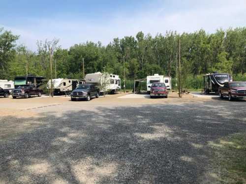 A gravel parking area with several RVs and trucks surrounded by trees and greenery.