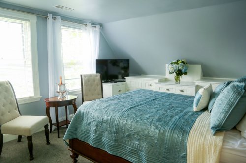 A cozy bedroom with a blue quilt, two chairs, a small table, and a TV, featuring natural light from large windows.