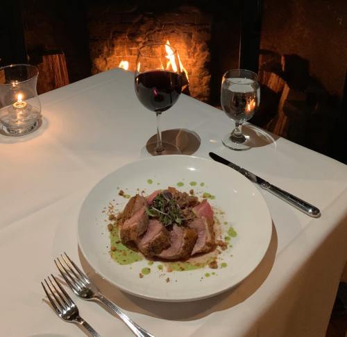 A beautifully plated dish of meat with sauce, accompanied by a glass of red wine, set against a cozy fireplace backdrop.