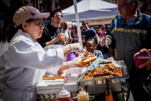 A vendor serves pulled pork on a bun to a customer at a busy outdoor food market.
