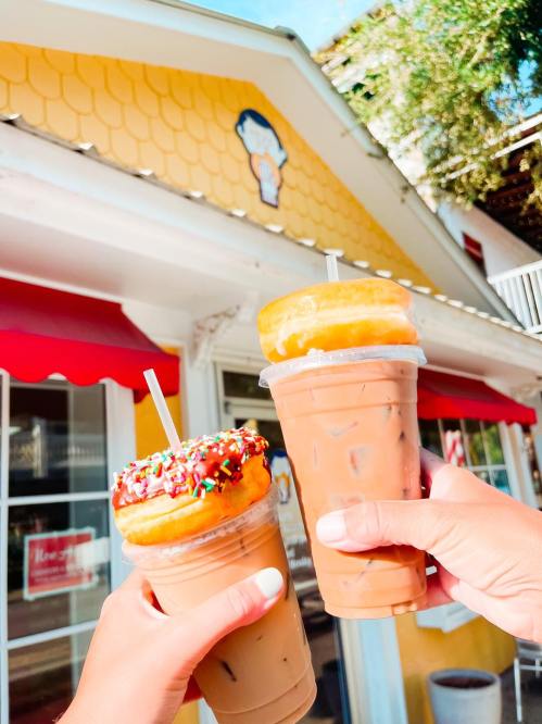 Two hands hold iced coffee drinks topped with donuts, in front of a bright yellow building with a cartoon logo.