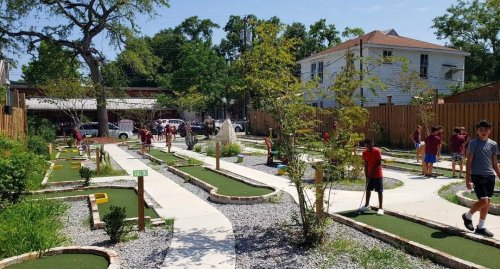 A mini-golf course with children playing, surrounded by greenery and buildings on a sunny day.