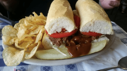 A sandwich with beef, tomatoes, and sauce on a hoagie roll, served with potato chips and a pickle on a plate.