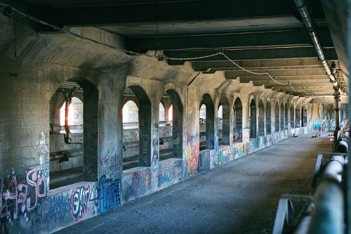 An empty, graffiti-covered underpass with arched supports and a dirt floor, illuminated by natural light.