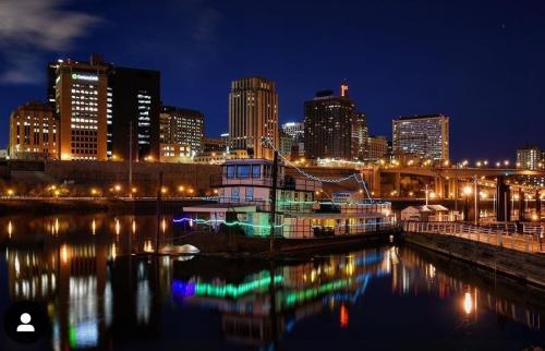 A nighttime cityscape featuring a lit boat on the water, with skyscrapers reflecting in the river.