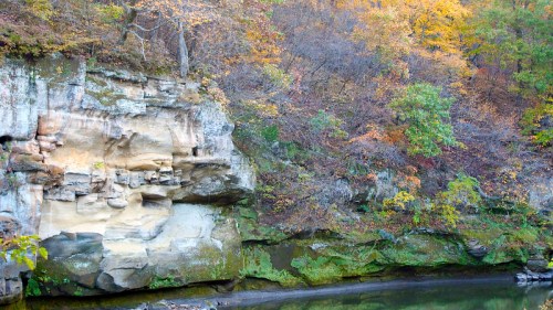 A rocky cliff with colorful autumn foliage reflected in a calm body of water below.
