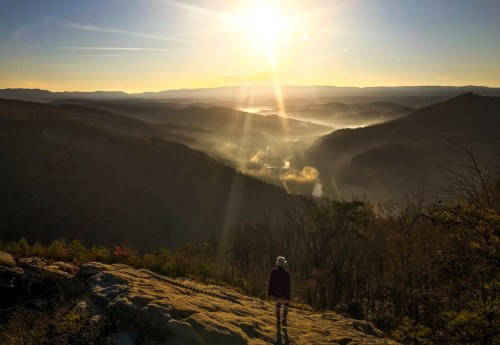 A person stands on a rocky ledge, overlooking a misty valley at sunrise, with rays of sunlight illuminating the landscape.
