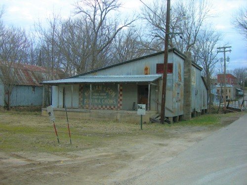 Abandoned building with a weathered sign, surrounded by bare trees and a dirt road.