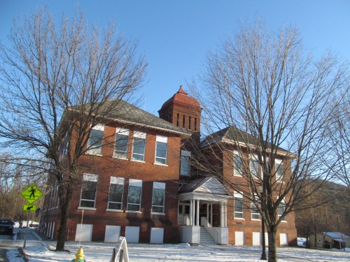 Historic brick building with a tower, surrounded by bare trees and a clear blue sky, in a snowy landscape.