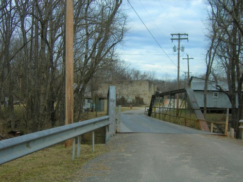 A rural scene featuring a metal bridge over a creek, surrounded by bare trees and a cloudy sky.